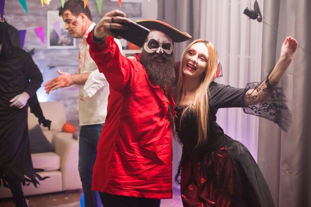 Mulher vampira feliz e homem pirata tirando uma selfie na celebração do halloween.