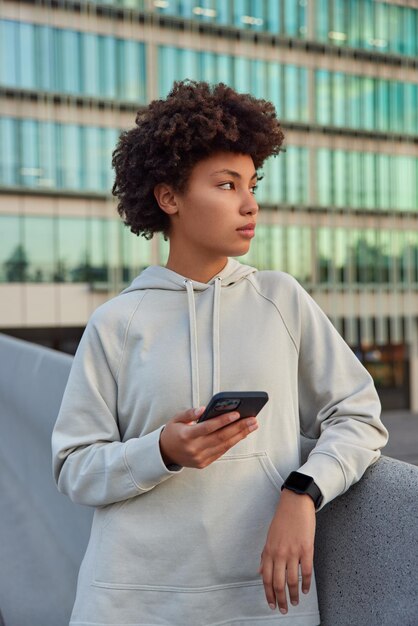 mulher vestida com capuz descansa em local urbano após a sessão de treino usa gadget moderno para navegar no site e a publicação parece estar perdida em pensamentos