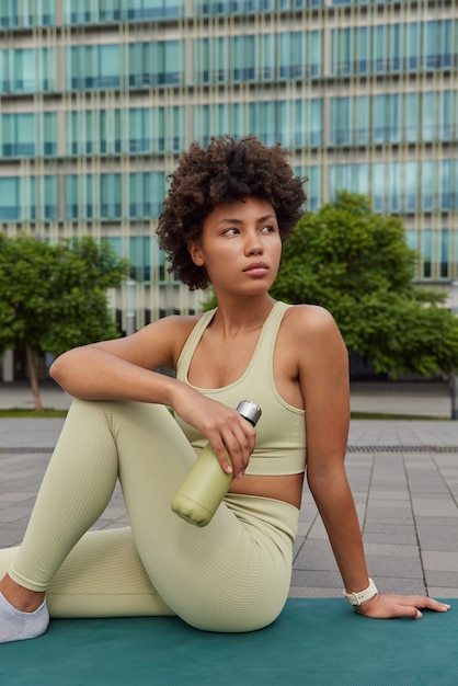 mulher vestida com roupas esportivas faz uma pausa após a sessão de treino sentada no karemat bebidas água segura garrafa desviando o olhar poses em ambiente urbano