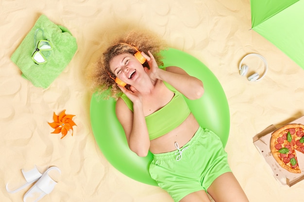 mulher vestida com top cortado e shorts gosta de ouvir música através de poses de fones de ouvido na praia sozinha come pizza encontra-se na pista de natação inflada verde.