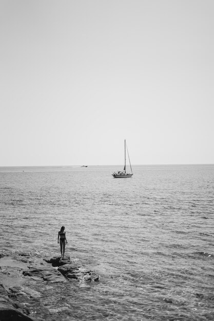 Mulher vestindo biquíni em pé em uma pedra pelo corpo de água com um veleiro flutuando no mar