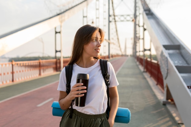 Mulher viajando com mochila posando na ponte enquanto segura a garrafa térmica