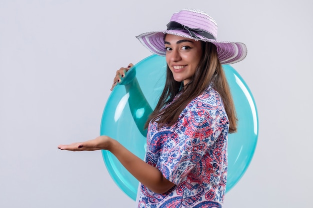 Foto grátis mulher viajante com chapéu de verão segurando um anel inflável positivo e feliz sorrindo alegremente apresentando com o braço da mão algo em pé no branco isolado