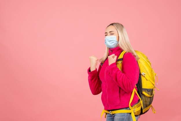 Mulher viajante com mochila amarela e máscara mostrando gesto vencedor com os olhos fechados