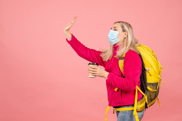 Mulher viajante com mochila amarela usando máscara segurando copo chamando alguém