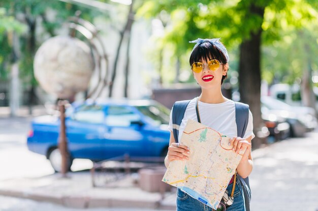 Mulher viajante feliz com cabelo preto curto explorando novas terras, segurando o mapa da cidade e sorrindo