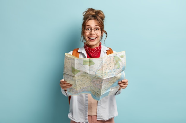 Mulher viajante feliz segura mapa, verifica rota para destino, viaja pela Europa durante as férias, carrega mochila, usa óculos redondos