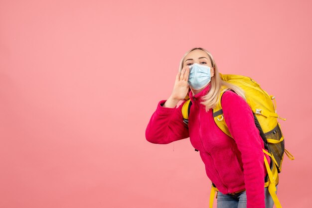 Mulher viajante fofa com mochila amarela e máscara chamando alguém de frente