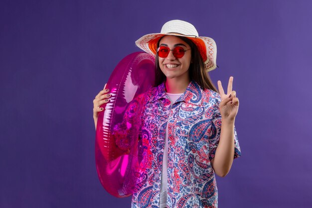 mulher viajante usando chapéu de verão e óculos de sol vermelhos segurando um anel inflável com uma cara feliz sorrindo alegremente fazendo a vitória cantar em pé no roxo