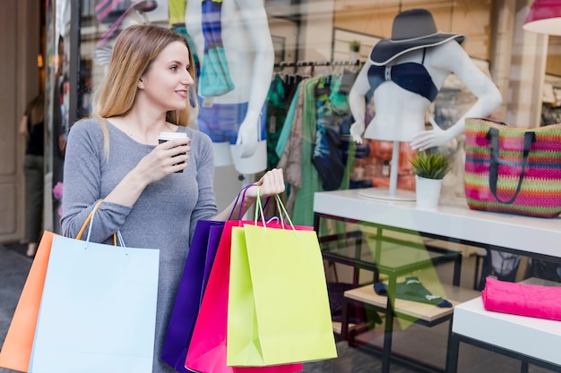 Mulher viciada em compras com bebida olhando t vitrine de uma loja