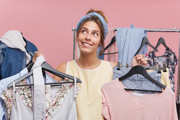 Mulher viciada em compras estar na boutique, escolhendo muitas roupas, olhando com expressão sonhadora, sem saber que roupa escolher para sair com o namorado. Alegre comprador feminino de roupas da moda