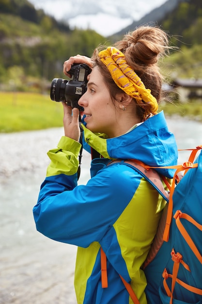 Mulher Wanderlust caminha em prados verdes nas montanhas, tira fotos maravilhosas com câmera digital, aprecia a beleza da paisagem natural, usa jaqueta