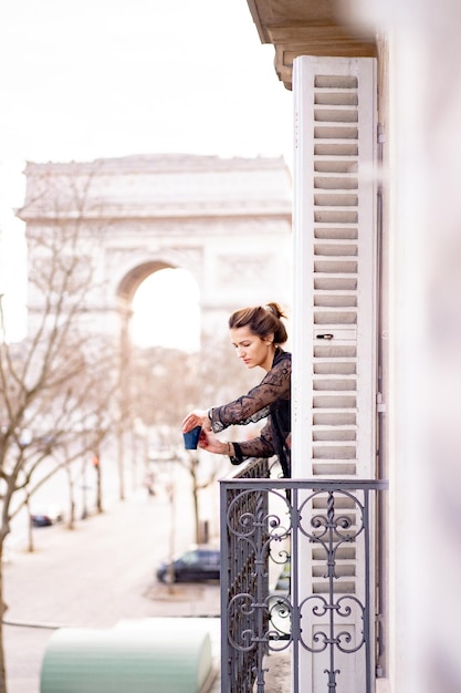 Mulher yang atraente de pijama está bebendo café na varanda de manhã na cidade de paris. vista do arco triunfal.