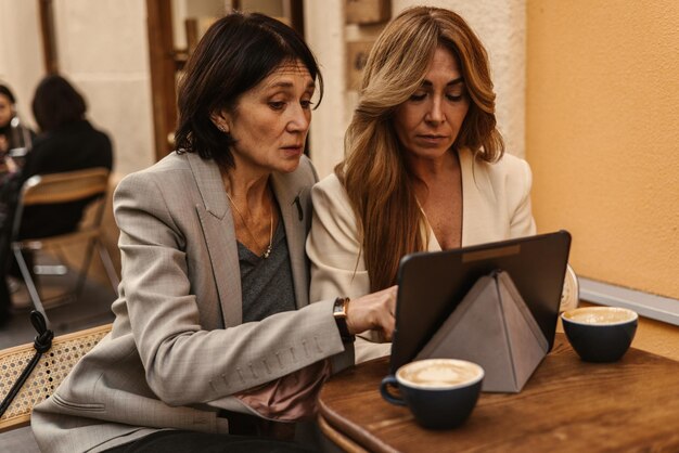 Foto grátis mulheres adultas caucasianas focadas trabalham em tablet enquanto estão sentadas no café de rua com uma xícara de café conceito de dispositivo tecnológico