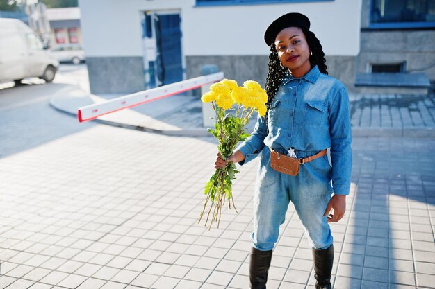 Mulheres afro-americanas elegantes em jeans e boina preta com buquê de flores amarelas posadas ao ar livre em dia ensolarado contra o edifício moderno azul