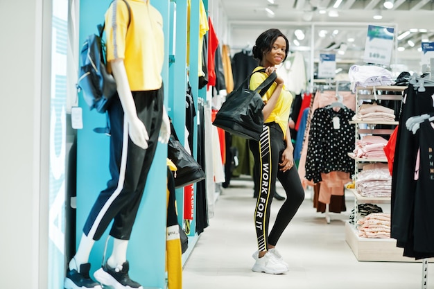Mulheres afro-americanas em agasalhos comprando no shopping de roupas esportivas com bolsa esportiva contra prateleiras Tema da loja esportiva
