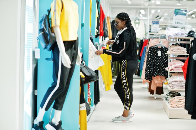 Mulheres afro-americanas em agasalhos e óculos de sol fazendo compras no shopping de roupas esportivas contra prateleiras Ela escolhe camiseta amarela Tema da loja de esportes