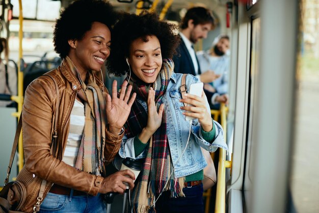 Mulheres afro-americanas felizes tendo chamada de vídeo por telefone inteligente enquanto se deslocam em um transporte público