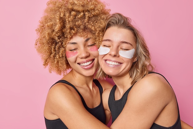 Foto grátis mulheres alegres e amigáveis se abraçam com sorriso de amor, aplicam suavemente manchas de beleza sob os olhos para tratamento e rejuvenescimento da pele, vestidas com camisetas pretas isoladas sobre fundo rosa