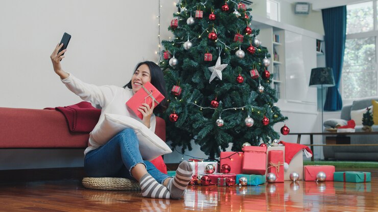 Foto grátis mulheres asiáticas comemoram o festival de natal. adolescente feminino relaxar feliz segurando presente e usando o smartphone selfie com árvore de natal desfrutar de férias de inverno natal na sala de estar em casa.