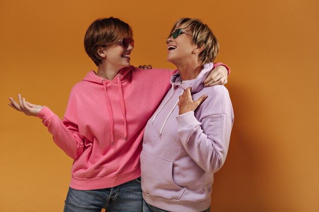Mulheres de cabelos curtos com óculos de sol em modernos moletons lilás e rosa com capuz e jeans, sorrindo e se abraçando em um pano de fundo laranja isolado.