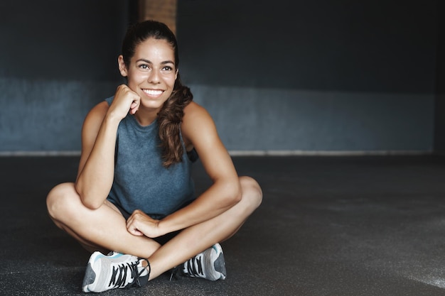 Mulheres de resistência e conceito de fitness bonita jovem atleta feminina saudável e esportiva termina o treinamento produtivo respire após o exercício sente-se com as pernas cruzadas no chão da academia sorrindo satisfeito