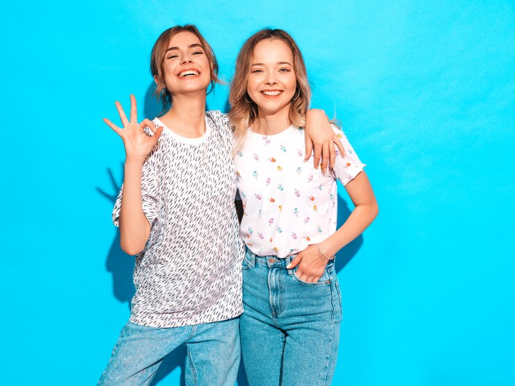 Foto grátis mulheres despreocupadas "sexy" que levantam perto da parede azul. modelos positivos se divertindo. mostra o sinal de ok