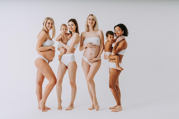 Mulheres grávidas felizes com barriga grande e lindas mães com bebês em estúdio - grupo multirracial de mulheres grávidas e mães com crianças vestindo roupas íntimas - gravidez, maternidade, pessoas, postura corporal Foto Premium