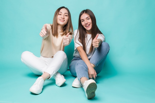 Mulheres jovens animadas com polegares para cima, posando no chão na parede turquesa.