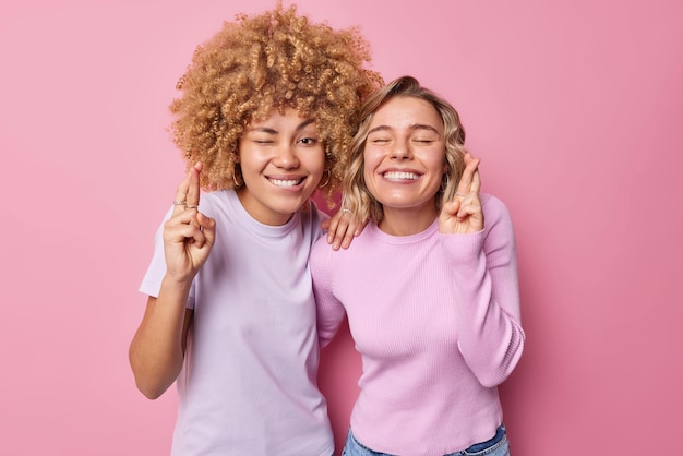 Mulheres jovens felizes positivas ficam esperançosas piscando os olhos mordem os lábios e mantêm os dedos cruzados acreditam na boa sorte vestida casualmente isolada sobre fundo rosa Dois alunos aguardam os resultados do exame