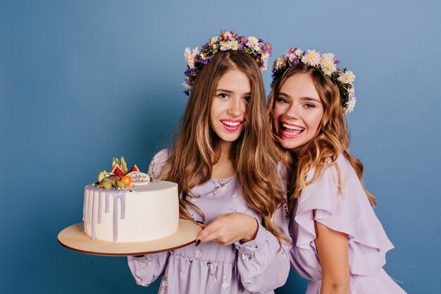 Mulheres lindas em vestidos roxos em pé na parede azul com um grande bolo cremoso