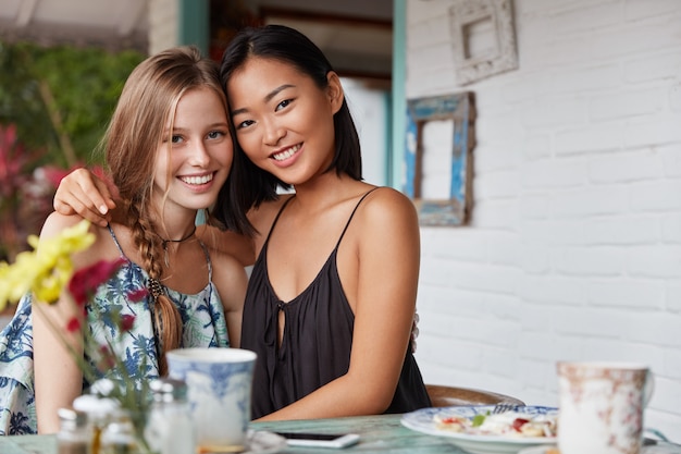 Mulheres mestiças bonitas de diferentes nacionalidades abraçam como demonstram amizade ou relacionamentos inter-raciais verdadeiros, bebem café no bar