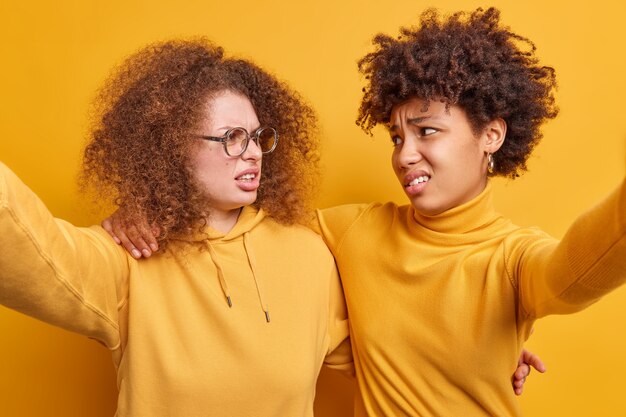 Mulheres mestiças insatisfeitas, rostos sorridentes, olham tristes uma para a outra, se abraçam e fazem selfie alguns problemas, vestir roupas casuais isoladas sobre uma parede amarela. Reações humanas negativas