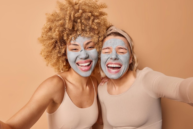 Mulheres muito felizes riem alegremente posam uma ao lado da outra se divertem fazer foto selfie manter os braços estendidos cuidar da pele usa máscaras de beleza de argila nutritivas isoladas sobre parede marrom conceito de beleza