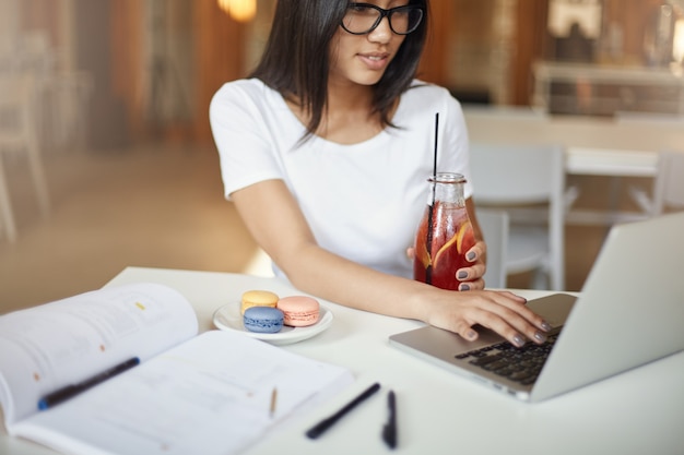 Mulheres são inteligentes. Jovem mulher usando um laptop, bebendo limonada ao mesmo tempo em um café, esperando para comer um bolo de macaron.
