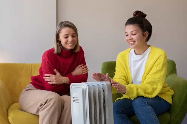 Mulheres sorridentes de tiro médio se aquecendo perto do aquecedor