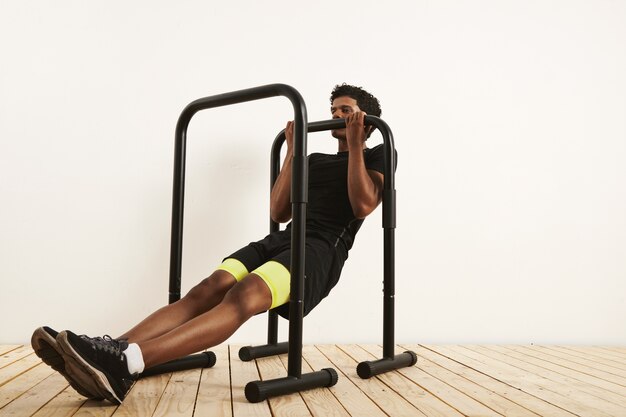 musculoso atleta afro-americano em roupa de treino preta, fazendo linhas de peso corporal em barras móveis contra uma parede branca e piso de madeira clara.