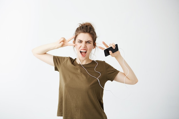 Música de escuta alegre bonita da mulher nos fones de ouvido que canta mostrando a paz sobre o fundo branco.