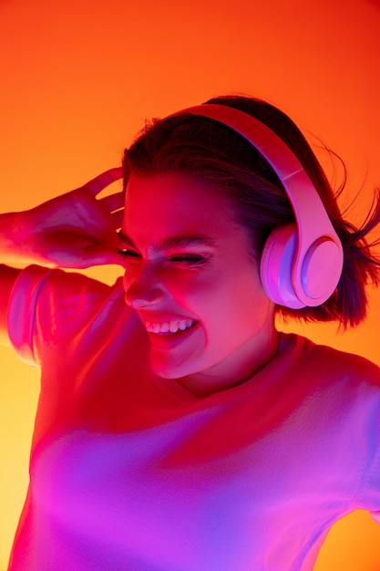 Música. Retrato de uma mulher caucasiana em fundo rosa studio em luz de néon da moda. Bela modelo feminino com fones de ouvido. Conceito de emoções humanas, expressão facial, vendas, anúncio, moda.