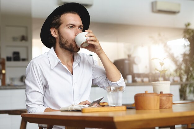 Na moda jovem com barba, chapéu e camisa branca com bebida quente, sentado à mesa e segurando o gadget na mão. Homem caucasiano, usando telefone celular, bebendo chá ou café no café acolhedor
