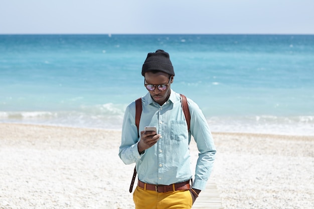 Na moda jovem turista masculino americano africano usando telefone celular na praia deserta, postando fotos da bela vista do mar ao seu redor através de mídias sociais com oceano azul e céu azul no horizonte