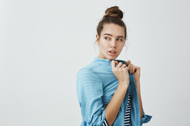 Na moda mulher escolhe roupas na loja. Foto de estúdio de namorada atraente feminina, puxando a camisa e olhando de lado em pé meio virado, tentando encontrar roupa combinando