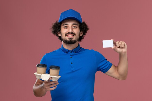 Na parede rosa, vista frontal do mensageiro masculino de uniforme azul e boné com xícaras de café para entrega e cartão nas mãos