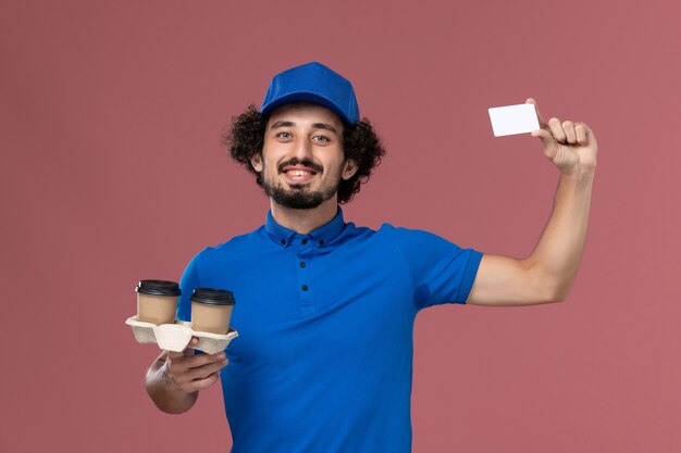 Na parede rosa, vista frontal do mensageiro masculino de uniforme azul e boné com xícaras de café para entrega e cartão nas mãos