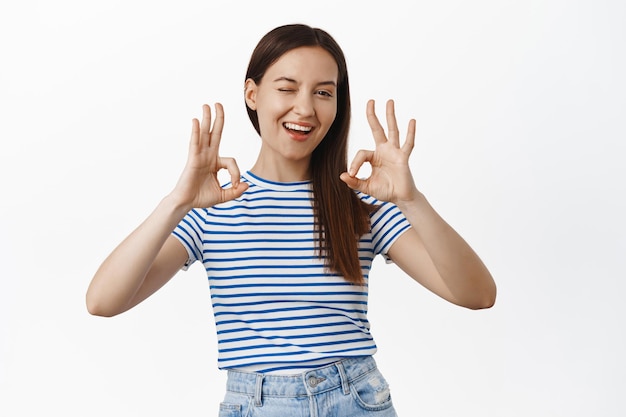 Nada mal, muito bem. Sorrindo alegre menina de 20 anos, piscando e mostrando sinal de OK OK, encorajá-lo, garantir tudo sob controle, tudo está bem perfeito, satisfeito contra o fundo branco.