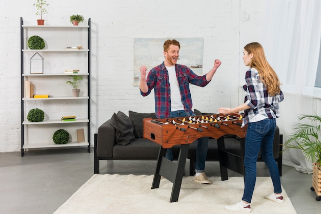 Namorada encolher os ombros na frente do homem celebrando seu sucesso perto do futebol de mesa em casa