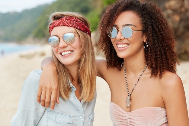 Foto grátis namoradas jovens têm expressões positivas de felicidade, se abraçam na praia, têm relacionamento inter-racial. mulher afro de pele escura jovem sorridente admirar o pôr do sol juntos ao ar livre casal homossexual romântico