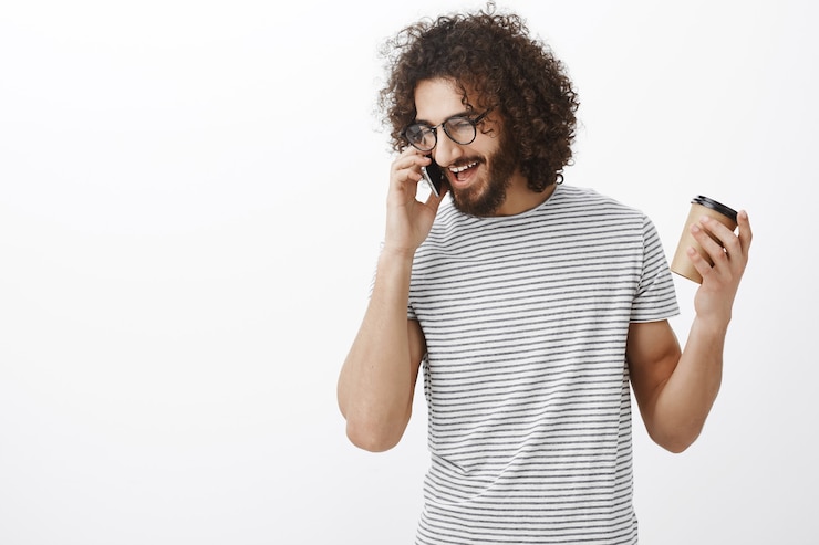 Foto grátis namorado adulto feliz satisfeito com cabelo encaracolado, usando óculos e uma elegante camiseta listrada, segurando a xícara e gesticulando enquanto fala no smartphone