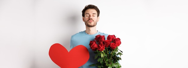 Foto grátis namorado romântico esperando beijo segurando buquê de flores rosas e grande coração vermelho no dia dos namorados