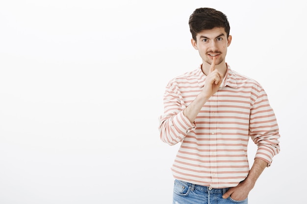 Não diga mais. Retrato de um homem europeu confiante interessado com barba e bigode, fazendo gesto de shush, dizendo shh com o dedo indicador sobre a boca, sorrindo e pedindo para manter a voz baixa sobre a parede cinza